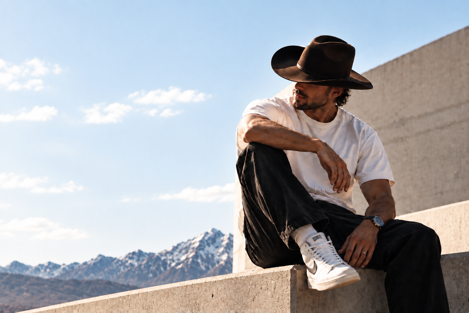 A man in a dark Western hat and modern streetwear seated against concrete architecture with the Wasatch mountains behind him